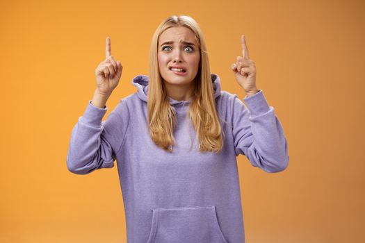 Worried perplexed unsure cute blond woman presenting project standing awkward afraid humiliation crining scared pointing up raised index finger waiting reply nervously, orange background