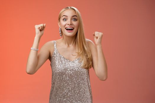 Surprised happy celebrating blond young woman in silver trendy dress raising hands up yes victory gesture smiling broadly excited winning first place reach goal grinning thrilled triumphing