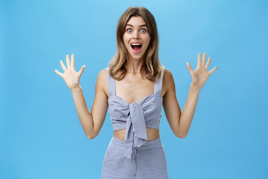 Cute woman with gapped teeth feeling exciting from surprise and anticipation smiling broadly raising palms from thrill and amazement popping eyes at camera posing over blue background