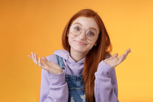 Nothing much. Attractive silly unsure carefree redhead unbothered cute female wearing geek glasses smirking uncertain shrugging hands spread sidways clueless what do future, orange background