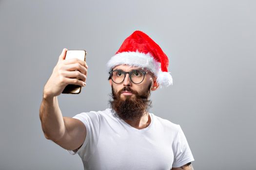 Christmas, holidays, technology and people concept - handsome bearded man in santa hat taking selfie picture with smartphone on grey background
