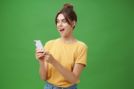 Woman reading surprising satisfying message in smartphone opening mouth from excitement, smiling amazed looking astonished at cellphone screen posing against green background in casual yellow t-shirt