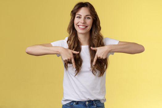 Lively helpful good-looking caucasian girl curly long hair showing you excellent place find hairdresser pointing down index fingers smiling toothy happy delighted grin stand yellow background