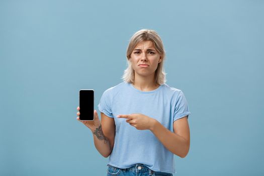 Unhappy sad attractive silly girl with fair hair tattoos and tanned skin frowning with regret and disappointment poonting at smartphone screen as if being dissatisfied with new girlfriend of ex-lover