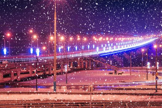 Traces of headlights from cars moving at winter night on the bridge, illuminated by lanterns in a snowfall. Lights reflecting in the wet asphalt.