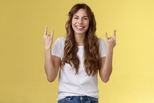 Happy cheerful feminine caucasian girl curly hairstyle show rock-n-roll heavy metal gesture smiling broadly enjoy awesome concert atmosphere stand yellow background joyfully cheering satisfied