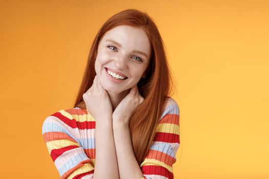 Silly enthusiastic attractive redhead blue-eyed girl tilting head touching neck flirty smiling enjoy perfect day feel happiness joy giggling coquettish flirting boyfriend, standing orange background