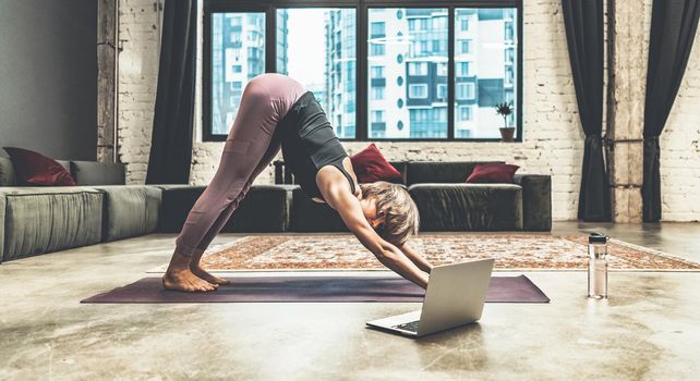 Nice Woman Practicing Yoga, Doing Down Dog Exercise, Watching Online Yoga Lessons by her Laptop, Yoga at Home. Close-up. Multi-storey Residential Building Background
