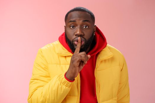 Waist-up serious-looking african-american bearded man in yellow jacket red hoodie bend camera asking keep quiet say shhh show shush gesture index finger on lips, confidential information taboo