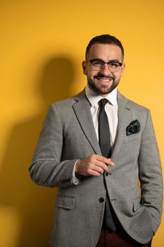 Handsome bearded smiling elegant man wearing eye glasses in formal wear with one hand lifted standing isolated on yellow background