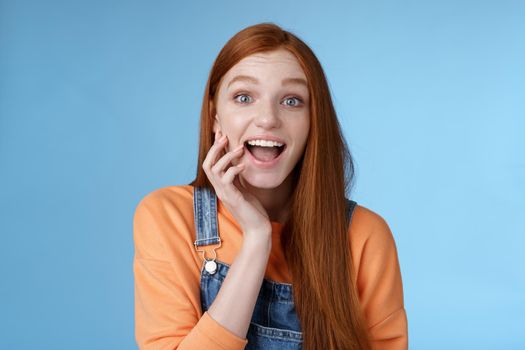 Excited enthusiasitc charismatic redhead female friend look surprised amused touch cheek open mouth thrilled discussing future prom rejoicing standing blue background joyful delighted