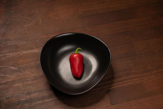 Organic mini red bell pepper laying lonely in a deep black bawl and ready to eat isolated on wooden background