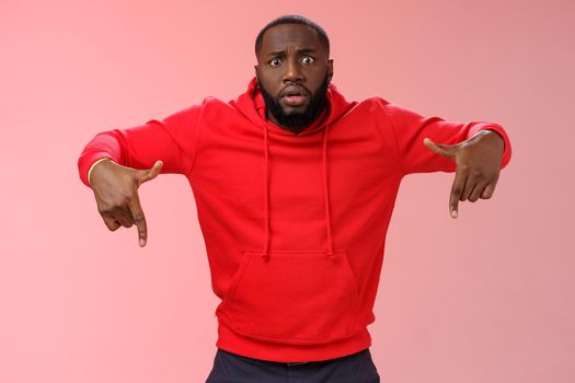 Nervous upset gasping young bearded african american man widen eyes shocked pointing down lose hope, standing worried anxious losing pari, wearing red hoodie pink background