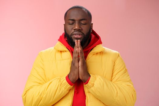 Serious-looking faithful young determined african-american praying guy in yellow coat press palms together pray close eyes whisper supplication asking god help make wish, standing pink background