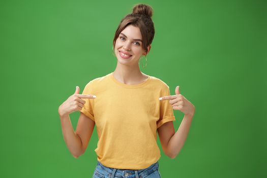 Calm and polite friendly-looking skillful young female coworker in yellow t-shirt pointing at herself self-assured tilting head and smiling at camera wanting participate in event over green background