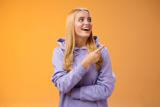Amused carefree joyful blond european woman in hoodie pointing looking right pleased smiling happily enjoying interesting fascinating peromance making choice in store shopping, orange background