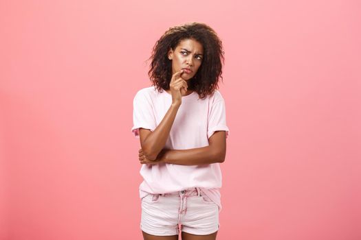 Suspicious displeased dark-skinned young curly-haired woman in trendy t-shirt and shorts saying hmm touching chin with finger frowning and staring left doubtful and unsure having concern