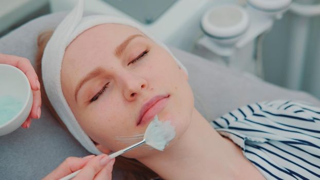 Cosmetician hands applying cream mask on young woman's face at beauty spa salon