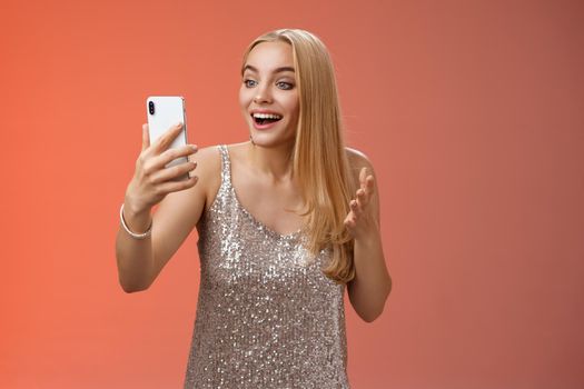Charming elegant nice blond girl in silver dress talking video call speaking looking smartphone display amused surprised smiling happily have conversation sibling showing prom outfit