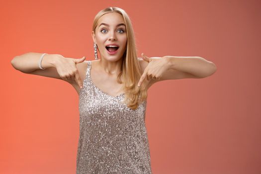 Impressed excited attractive glamour blond girl in silver glittering dress gasping thrilled pointing down glance camera fascinated check out fabulous awesome promo, standing amused red background