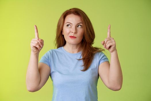 Curious thoughtful cute redhead female middle-aged ginger mother smirking thinking look aside pointing up index fingers deciding choice pondering choosing between variants green background