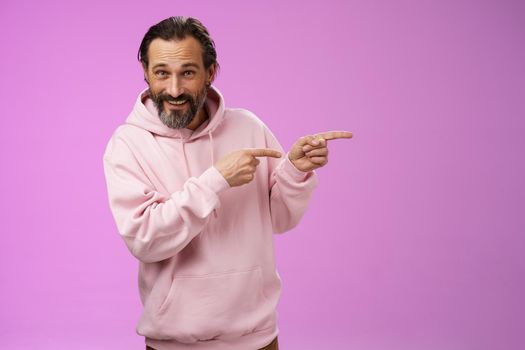 Sassy confident good-looking macho man 50s in pink hoodie smiling broadly inviting join pointing right showing interesting amusing place hang out welcoming take look, standing purple background