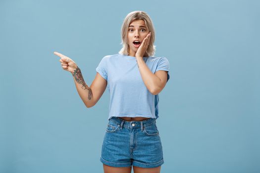 Waist-up shot of worried surprised emotive blonde woman in t-shirt saying gosh opening mouth from shock holding palm on cheek looking with empathy while pointing left with tattooed arm over blue wall