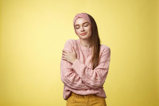 Sweet glamour young girl wearing warm comfy sweater embracing herself crossing arms over body in hug, smiling soft and kind close eyes, recalling lovely days, tender memories over yellow wall