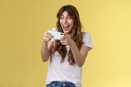 Happy upbeat playful enthusiastic curly-haired girl tempting playing stunning awesome smartphone game hold mobile phone horizontal way smiling broadly stare camera focused winning beat score