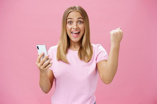 Girl beat own record in smartphone game raising clenched fist in cheer and triumph holding cellphone, smiling excited and happy at camera celebrating victory with joyful gesture over pink background