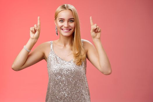 Excited gorgeous blond european woman in silver shiny elegant dress raise hands pointing up showing impressive incredible advertisement smiling happily thrilled wanna take closer look, red background