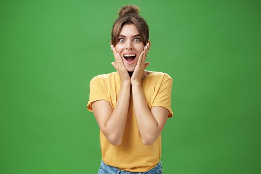 Indoor shot of excited and surprised energized attractive glamorous female in yellow t-shirt touching cheeks from amazement smiling broadly astonished posing over green background