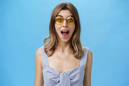 Waist-up shot of excited surprised and emotive charismatic caucasian woman in trendy sunglasses opening mouth from amazement and joy gazing at camera impressed and astonished over blue background