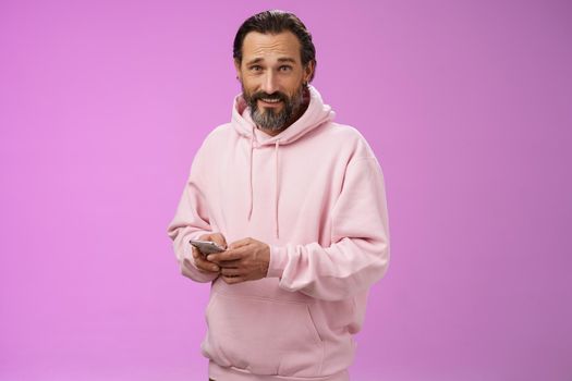 Stylish handsome adult bearded guy 40s in trendy pink hoodie holding smartphone texting look camera unbothered cool confident distracted playing awesome new app game, purple background