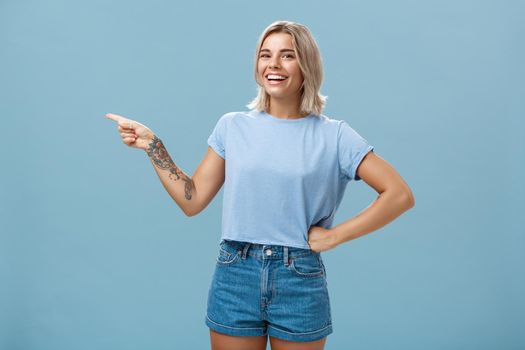 Girl proudly shows place where she works holding hand on waist in confident pose smiling and laughing happily pointing left with tattooed arm standing over blue background carefree