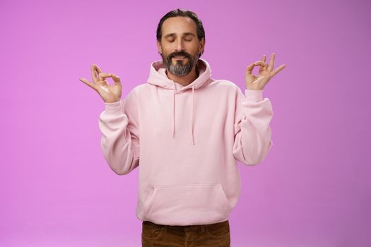 Peaceful charming hipster old man wearing cool pink hoodie close eyes breathing practice release stress meditating standing relaxed happy yoga meditation pose nirvana gesture, purple background