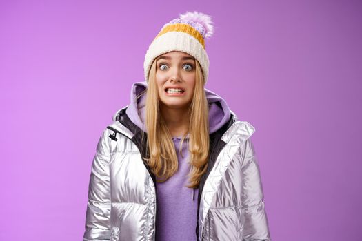 Awkward worried cute timid blond girl in silver jacket hoodie winter hat clench teeth popping eyes camera ooops make mistake standing nervous someone notice, purple background