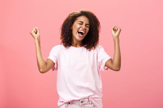 Amazed and happy triumphing african american sportswoman celebrating victory yelling from awesome exciting feelings closing eyes raising fists hight in win gesture standing over pink background