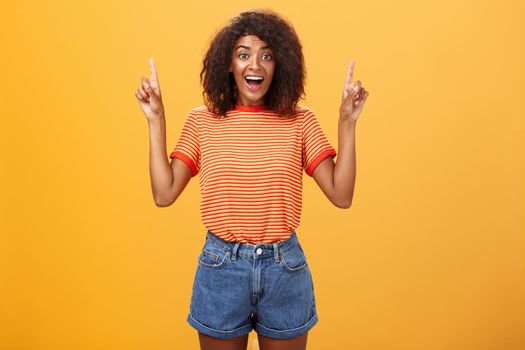 Charmed and impressed african american stylish woman seeing incredible and awesome item on sale raising hands and pointing up with broad grin being excited of new purchase over orange wall