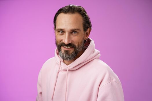 Charming alluring handsome bearded stylish adult male model earring pink hoodie smiling delighted express confidence positivity feel lucky amused, standing purple background talking casually