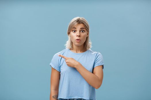 Studio shot of amazed stunned cute blonde witnessing unbelievable event gasping opening mouth staring astonished and pointing left being questioned and shocked over blue background