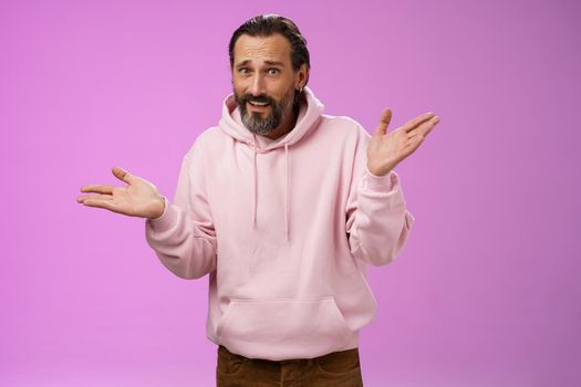 So what bite me. Portrait ignorant careless cool stylish mature bearded man earring pink hoodie shrugging hands sideways mocking being rude standing pissed unwilling help standing purple background