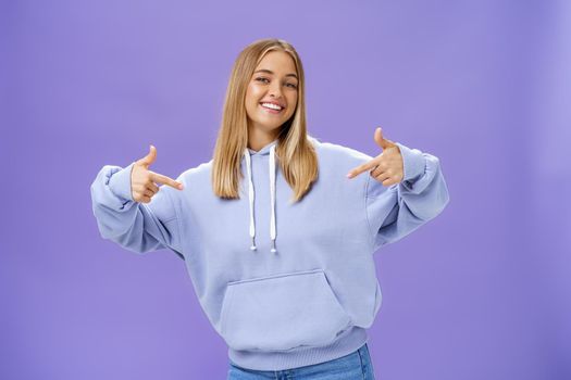 Self-assured cheerful carefree young girl with tanned skin and fair hair pointing proudly at herself bragging about own skills and achievements standing cool feeling awesome in new hoodie