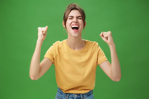 Waist-up shot of charismatic energized and excited female in yellow t-shirt closing eyes yelling from joy and happiness raising hands in cheer celebrating successful news over green background
