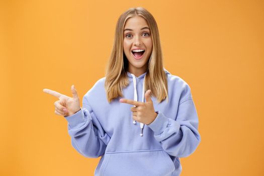 Amused girl pointing at curious copy space showing left with index fingers smiling excited and surprised standing entertained with upbeat grin in cozy blue hoodie over orange background