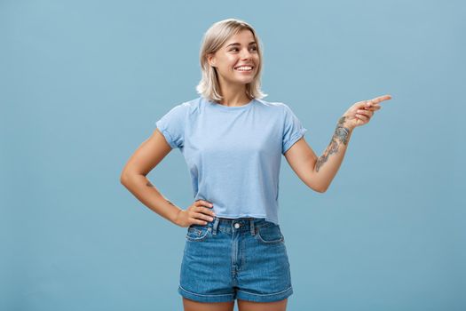 Bossy confident female manager with tattoo on arm holding hand on waist pointing and gazing right with pleased relaxed look giving directions to employees posing over blue background