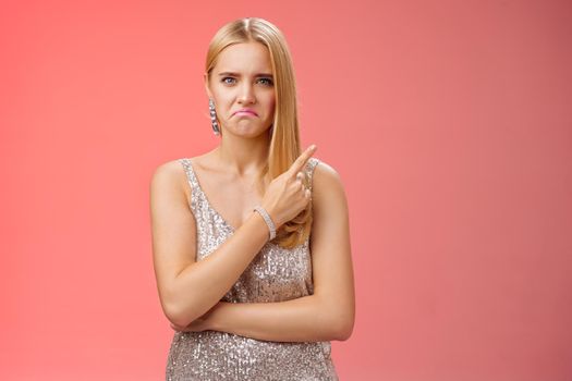 Jealous displeased angry young revengeful blond ex-girlfriend in luxurious silver shiny dress frowning pouting pointing upper right corner displeased pissed standing fed up upset red background