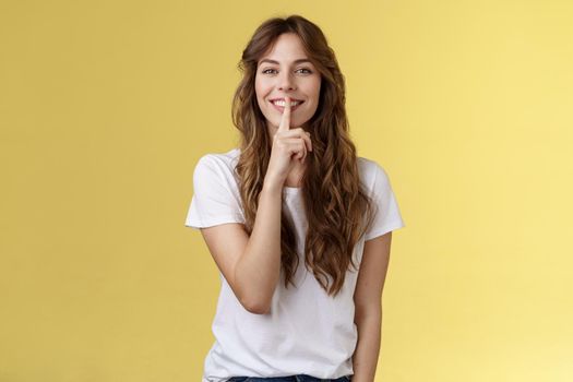 Cute cunning lovely european girl curly hairstyle hiding beauty secret smiling sensually show hush shush gesture index finger pressed lips grinning joyfully stand yellow background