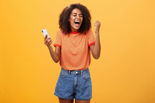 Portrait of ambitious happy young african american girl yelling from happiness and triumph clenching fist in joy and celebration feeling excited and relieved holding smartphone over orange wall