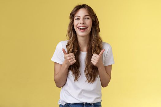 Pleased outgoing cheerful good-looking caucasian girl curly long haircut white t-shirt show thumbs up smiling lively pleased like awesome performance approve good choice yellow background